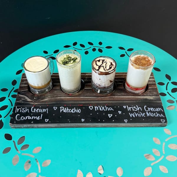 Four mini drinks, including Irish Cream, Pistachio, Mocha, and Irish Cream White Mocha, on a wooden tray.