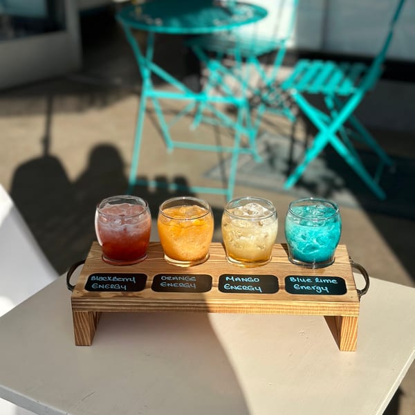 Four colorful energy drinks on a wooden tray, with outdoor seating in the background.