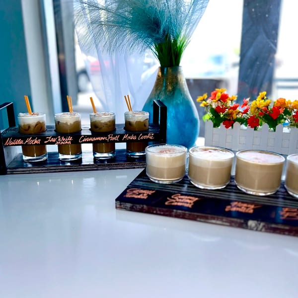 Two flights of coffee drinks, one with whipped cream, on a white table with flowers.