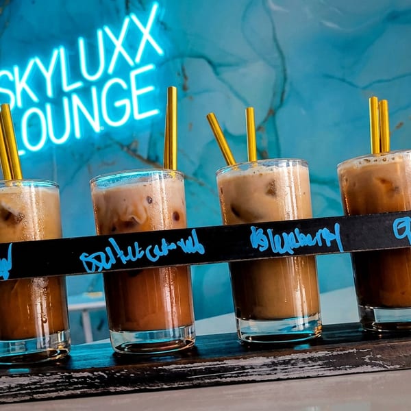 Four iced coffee drinks with golden straws in a wooden caddy, with a neon "SKYLUXX LOUNGE" sign.