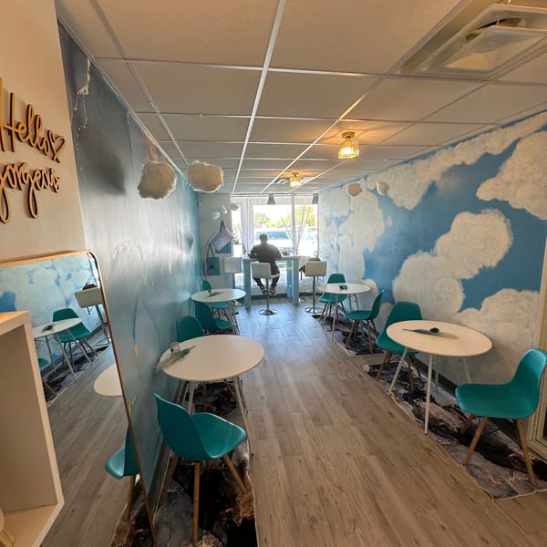 A cafe with cloud-painted walls, teal chairs, white tables, and a person at a high-top table.